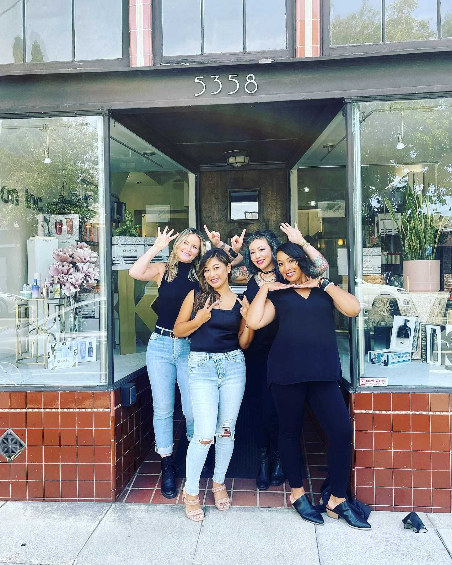 Group of four smiling women posing in front of a store with glass windows.