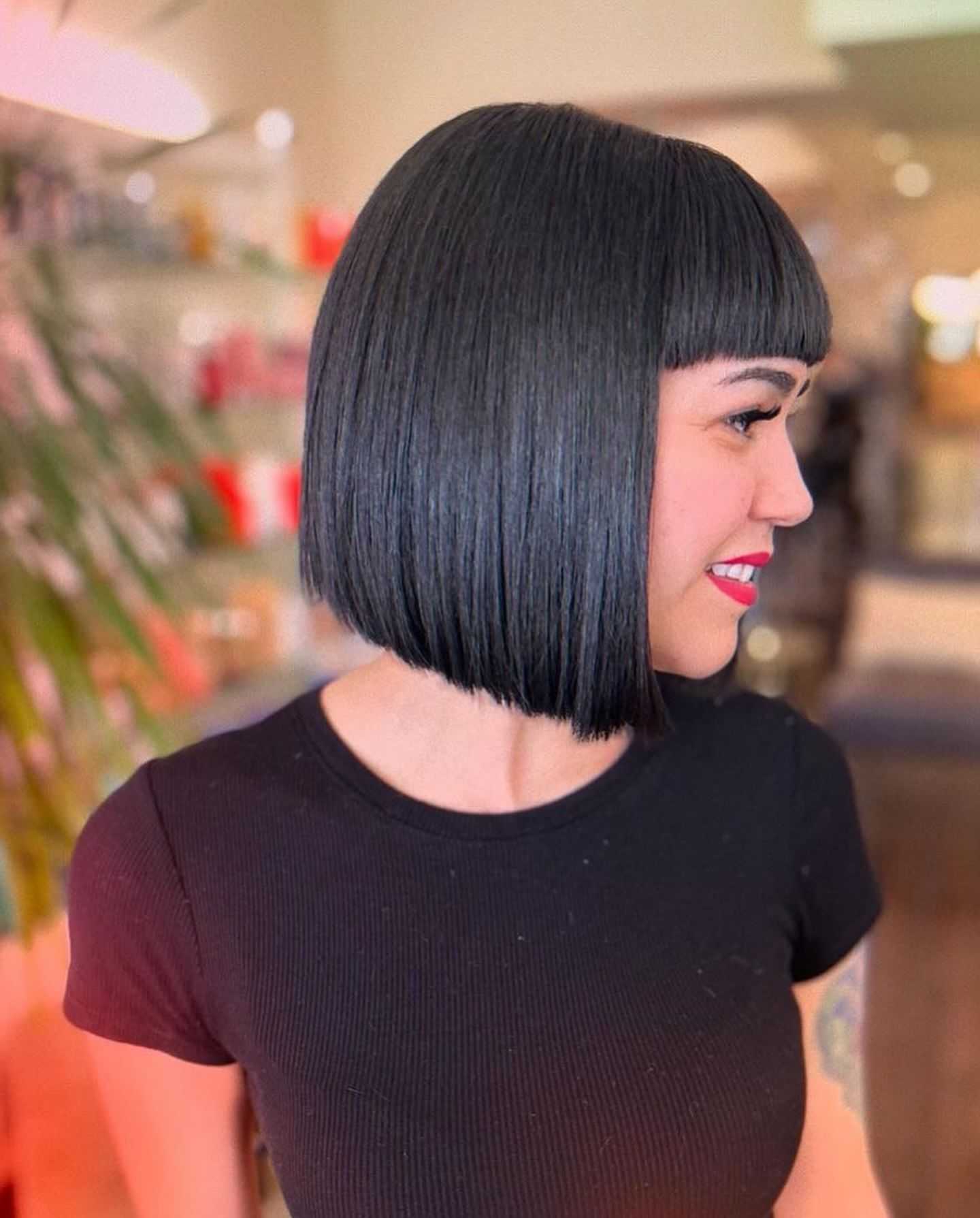 Woman with sleek black bob haircut and bangs, wearing a black shirt, smiling sideways.